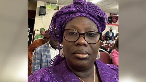 A selfie of a woman with black glasses and wearing a purple top and a purple headdress. She looks as though she's in a community centre with rows of chairs behind her