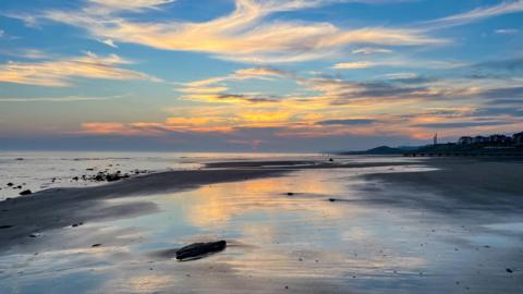 A beach at sunset. The sand is cast in shadow and the sky is blue at the top and moves into a golden colour towards the bottom. There are wispy clouds and the coastline in the distance is completely in shadow. 