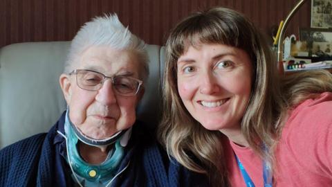 Hazel, on the right, a young woman with long brown hair, sat next to an elderly man on the left, who has a medical brace around his neck