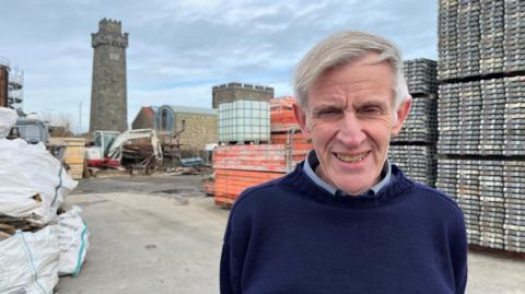 Smiling older man in navy jumper, known as a "Guernsey Knit", stands in a construction yard with scaffolding, machinery, and stone tower behind him under an overcast sky. 