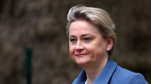 A close up of Yvette Cooper as she walks past some bushes. She is smiling slightly and wearing a blue jacket and white leaf-shaped earrings. Her hair is cropped short.