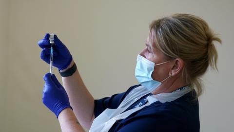 A female nurse is wearing blue scrubs and protective gear. She is wearing blue gloves and a face mask while holding a needle.