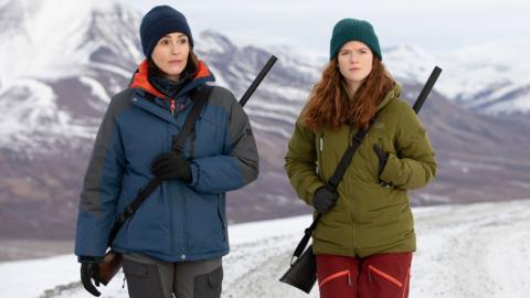 Suranna Jones as DCI Amy Silva and Rose Leslie as DI Kirsten Longacre walking through an icy landscape carrying guns on their back in Vigil