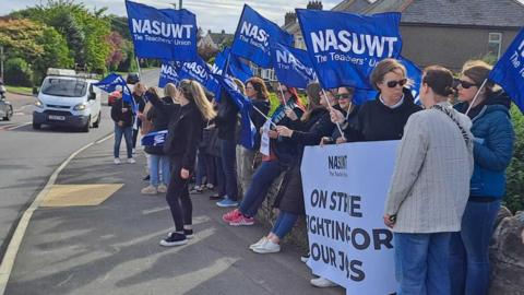 A group of people standing at the side of a road with blue NASUWT flags. Someone is holding a large, white sign which reads 'On strike fighting for our jobs'.