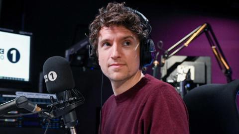 Greg James is wearing a burgundy jumper and is wearing headphones on his head. He is sitting at a desk in front of a microphone with a Radio 1 muff.