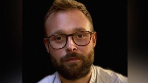 The head and shoulders of Matt Burgin-Powell against a dark background. He is wearing an Oxford-style blue cotton shirt, has a thick brown beard, short hair with a quiff, and glasses.