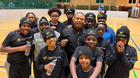 Gaby Kitoko, who is the centre of the picture and wearing a blue jacket with a silver zip, stands with just some of the youngsters who form part of the group. Most of them are wearing blue shirts that say 'Chance to Shine/Street' on them, and they are standing on a pitch they have just completed a game on.