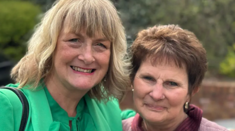 Ali France has blonde wavy shoulder-length hair and is wearing a bright green jacket and top. She has her arm round Sarah Forsyth, who has a nose ring and gold earring and has short brown hair.