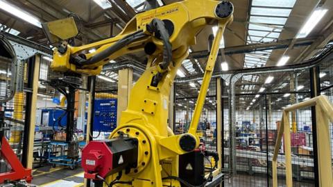 A robotic arm picker inside a factory. It is bright yellow with black parts and a black and red section at the front. It is a couple of metres tall and sits in front of mesh doors, in what looks like a warehouse or factory with overhead strip lighting and ceiling windows.