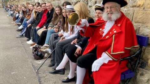A man in a red cloak holds a brass bell with a wooden handle. He has white whiskers and wears a black tricorn hat. He is sitting on a long railway bench on a railway platform. Next to him trailing into the distance sits another 238 people, some in period costume.