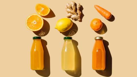 An orange, lemon, ginger, tangerine and a carrot above three bottles of orange and yellow liquid