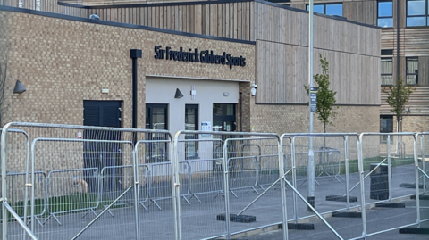 Metal heras fencing in front of a sports hall at the school, which is made of light-coloured bricks and has black lettering above a door that reads: "Sir Frederick Gibberd Sports".