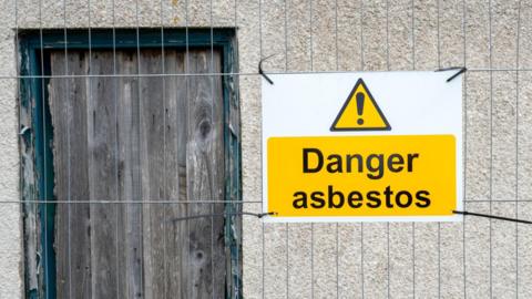 A yellow sign saying danger asbestos, tied to a metal fence