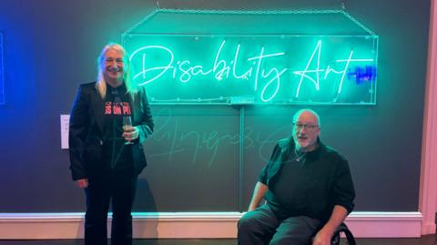 A woman with blonde hair is standing in front of a wall with a neon sign on it. She is wearing black trousers, suit jacket and t-shirt, which has pink writing on it. She is holding a champagne glass. There is a man next to her in a wheelchair. He is in all black and is wearing glasses, and is bald. The sign is illuminated in teal blue light, and reads "Disability Art"