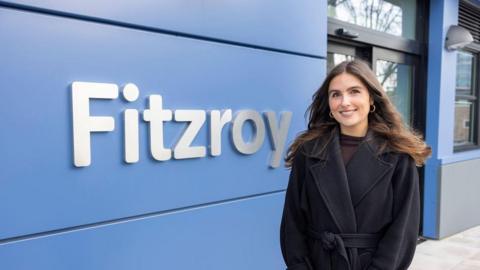 Shelley Oliver pictured outside the Fitzroy Building in a black coat. She has long brown hair and she is smiling. The building has a royal blue exterior with a large silver sign that reads Fitzroy.