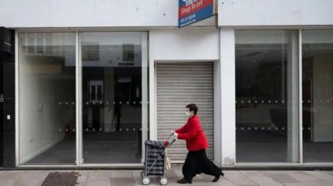 View of an empty shop front - with floor length windows and a roller over the door in the middle. A woman walks in front of it pushing a shopping trolley, and wearing a red jacked and black trousers.