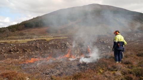Two men standing in the Scottish countryside as a fire burns in the brown grass