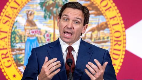 Ron DeSantis stands behind two microphones. He looks impassioned as he gives a speech. He's in front of a picture, surrounded by a golden border with the words "Great Seal of the State of Florida" written in red block capitals