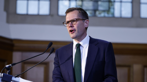 Reform UK's Robert Jenrick mid-speech. He is wearing glasses, has short, dark hair and is wearing a collared shirt and suit with a green tie.