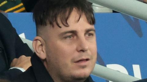 A man with messy black hair wearing a black coat. The image is zoomed in on his face, and he is looking off to the right of the screen