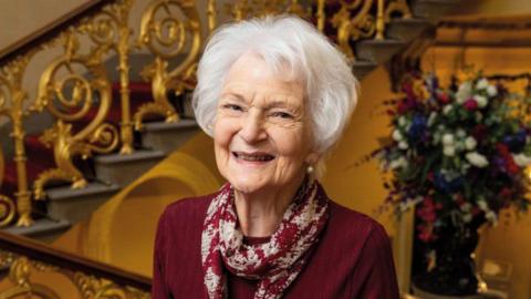 Dr Sheils Eith, wearing a red top and a white and red patterned scarf. She is looking at the camera while standing on a plush staircase.