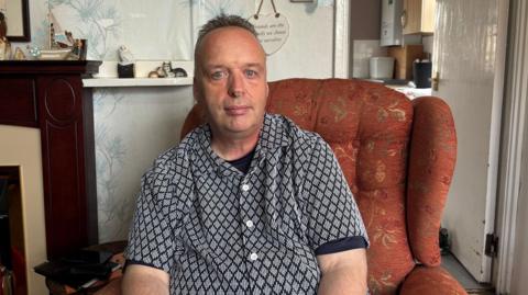 A man in an orange chair has dark hair swept back. He is wearing a black and white shirt