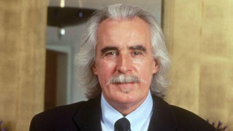 Charlie Miller has shoulder-length silvery hair and moustache. He is wearing a black suit jacket, white shirt and tie.