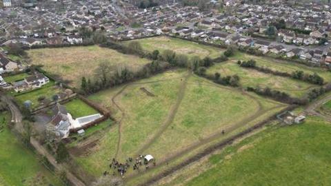 An aerial view of a several green fields with paths crossing them and hedges along the borders. A small crowd of people are standing in one corner of a field. In the distance, many houses can be seen, on the edge of Frome.