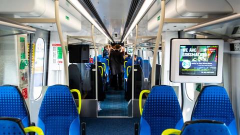 A view of a Northern train with blue seats and yellow handrails. We look down the middle of a carriage towards a number of passengers who are standing up in the distance.