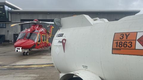 A white jet fuel tank is in the foreground with orange and black lettering, behind is the red and yellow Cornwall Air Ambulance parked outside the hangar