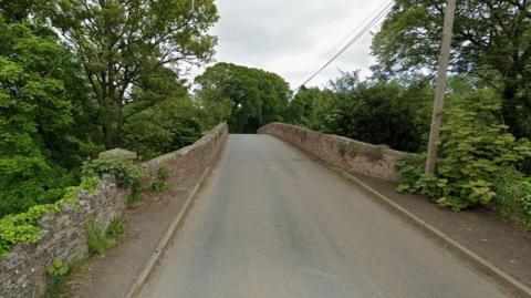 A Google street view image of a rural road going over a stone bridge. There is a telegraph pole on the right with wires stretching into the distance. Many trees surround the bridge and the walls of the bridge are covered in greenery. 