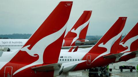 Four planes are lined up in a row on the tarmac of an aiport, each with the Qantas logo of a stylized white kangaroo on a red background emblazoned on the tail. 