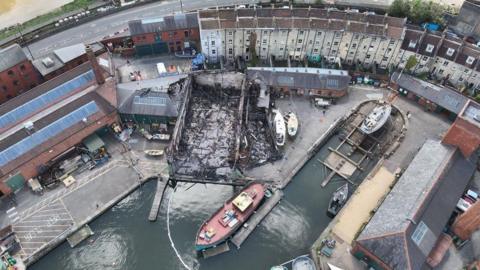An aerial image taken above the burnt shell of where the Underfall Yard buildings once stood. There is lots of charred debris and wood lying on the ground, the roof is gone and the inside has been gutted by fire.