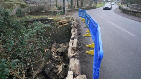 The damaged bridge in North Street, Midhurst, with the brick wall  demolished and a blue and yellow barrier fencing off the damaged section.