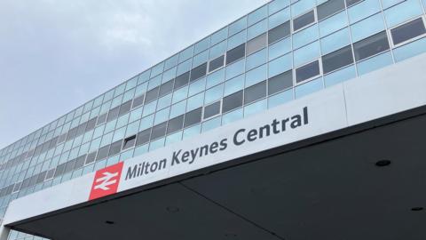 The outside of Milton Keynes railway station. There is a sign with the station name and glass windows above.