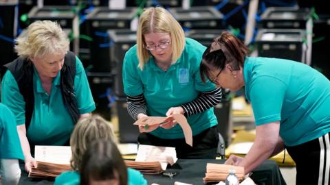 A group of vote counters wearing teal shirts, the colour of Perth council, start counting ballots on a table in Perth
