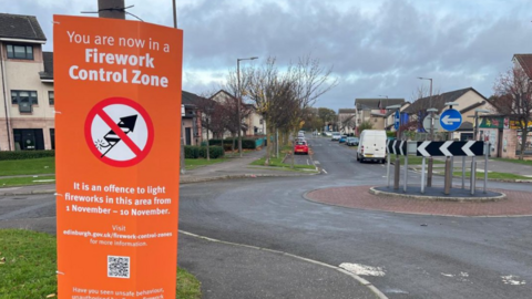 Orange sing tied to a lamp post saying firework control zone just in front of a roundabout with some houses in background