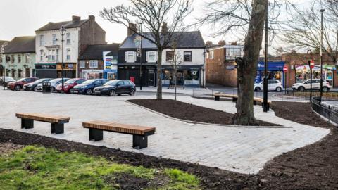 The pedestrian space at Harland Space. A paved area has been created near the high street. The pavement has been built around two trees. Fresh soil where grass and plants will be grown can be seen. Cars are parked by the road.