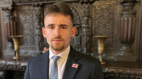 A brown-haired man with a goatee beard, wearing a navy suit, white shirt and blue tie, standing in front of a large carved wooden fireplace with with golden goblets
