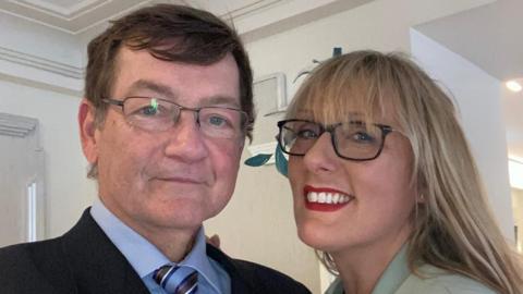 Phil and Juli. It is a selfie-style photo. Phil is waring a dark blazer, with blue shirt and stripy tie, Juli is wearing a green coloured top.