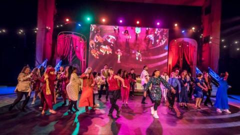 Cast members on stage dance in brightly coloured outfits under disco lights