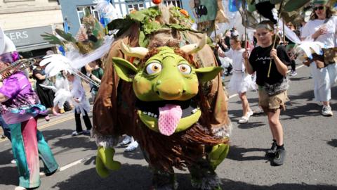 A green and brown carnival fantasy creature with horns and yellow eyes sticks an oversized pink tongue out at the camera. It is a sunny day with lots of other people dressed up for the carnival procession.