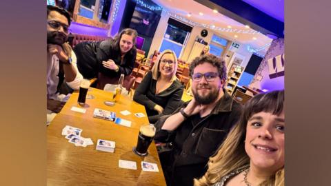 Five people - two men and three women - sat around a table in a pub. They are all smiling at the camera. Playing cards and drinks are on the table.