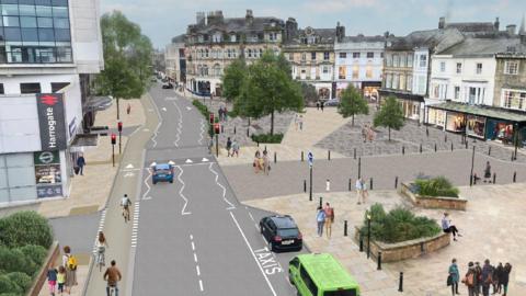 A digitally rendered view of a town centre with people walking and cycling along wide pavements and marked lanes, surrounded by shops and buildings, including one with a “Harrogate” sign.
