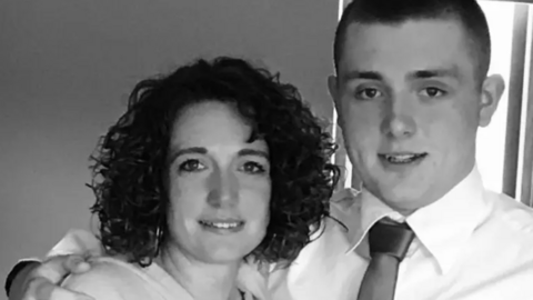 A black and white picture of Rachel Baker, and her son Joe. She has dark curly hair to her shoulders. He has short dark hair and wears a white shirt and dark tie.