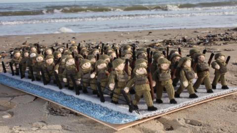 Small knitted soldiers placed in front of the sea on the beach.