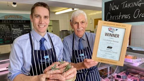 Norfolk butchers James Middleton and Richard Bowers pose in their shop with the award-winning Lincolnshire sausages and a certificate saying "Winner". They are wearing blue and white striped aprons and hold James, on the left of the picture, is holding a plate of raw sausages garnished with herbs.