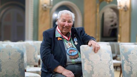 Justice Murray Sinclair sits in the ballroom at Rideau Hall, with his hand placed on a chair covered in a floral print in front of him. He is smiling at the camera and has grey hair. 