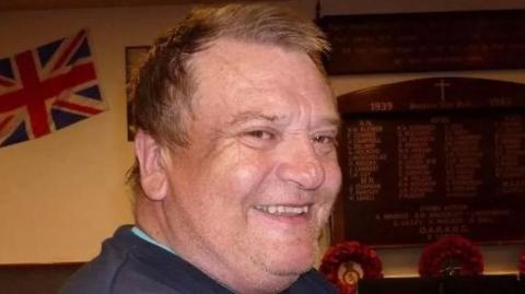 Peter Coates is looking over his shoulder and smiling brightly at the camera. He has short brown hair and is wearing a blue jumper. There is a wooden memorial board on the wall behind him, as well as a union jack flag.