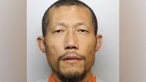 Hongchi Xiao looks at the camera with a blank expression in a custody picture. He is balding with dark hair and a dark goatee, and wears a grey sweatshirt over an orange top. 
